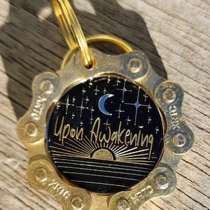 May include: A circular gold-toned metal tag featuring a bicycle chain link border. The center is a black disc with a silver crescent moon, stars, and a rising sun graphic. The text "Upon Awakening" is printed in gold script across the center.