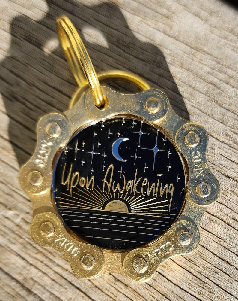 May include: A circular gold-toned metal tag featuring a bicycle chain link border. The center is a black disc with a silver crescent moon, stars, and a rising sun graphic. The text "Upon Awakening" is printed in gold script across the center.