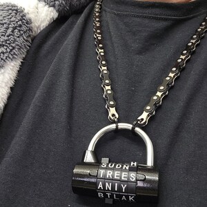 May include: A black necklace made of bicycle chain with a combination lock pendant. The lock displays the letters "SUDNH", "TREES", "ANIY", and "BTLAK". The necklace is worn over a black shirt and a gray and white patterned jacket.