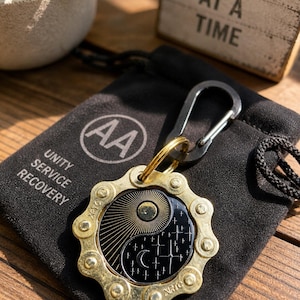 May include: A circular keychain featuring a brass-colored metal gear frame surrounding a black enamel inlay with a yin-yang design of a sun with rays and a moon with stars. The keychain includes a black carabiner and rests on a black fabric pouch.