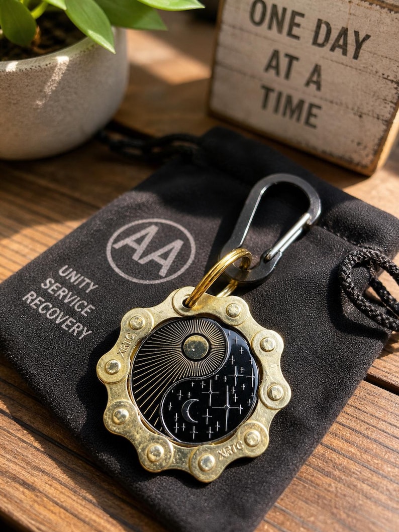May include: A circular keychain featuring a brass-colored metal gear frame surrounding a black enamel inlay with a yin-yang design of a sun with rays and a moon with stars. The keychain includes a black carabiner and rests on a black fabric pouch.