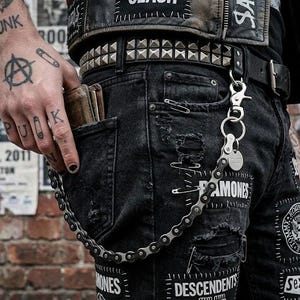 May include: Black denim jeans featuring distressed patches, safety pin embellishments, and various band patches including Ramones and Descendents. A metal bicycle chain wallet chain is attached to a studded leather belt with a silver-toned clasp.