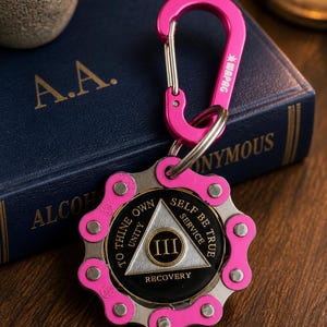 May include: A pink metal carabiner attached to a circular keychain featuring a bicycle chain link border. The centre medallion is black with a silver triangle containing the Roman numeral III, surrounded by the text TO THINE OWN SELF BE TRUE.
