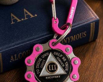 May include: A pink metal carabiner attached to a circular keychain featuring a bicycle chain link border. The center medallion is black with a silver triangle containing the Roman numeral III, surrounded by the text TO THINE OWN SELF BE TRUE.