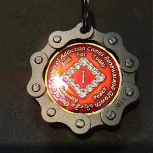 May include: A circular medallion keychain featuring a black metal bicycle chain border. The center is a red and gold coin with a diamond-shaped border of clear rhinestones, the Roman numeral I, and the text Out of the Ashes of Addiction Comes Renewal and Growth, Just For Today, Serenity, and Peace.
