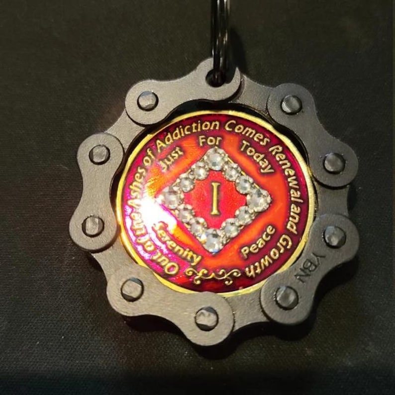 May include: A circular medallion keychain featuring a black metal bicycle chain border. The center is a red and gold coin with a diamond-shaped border of clear rhinestones, the Roman numeral I, and the text Out of the Ashes of Addiction Comes Renewal and Growth, Just For Today, Serenity, and Peace.