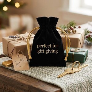 May include: A black velvet drawstring pouch with gold cords and the text "perfect for gift giving" printed in gold on the front. The pouch is positioned among various wrapped gift boxes, a small tag labeled "To Dad," and a gold-colored metal key.