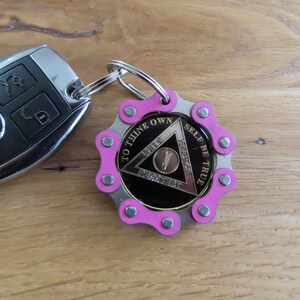May include: A black Mercedes car key fob attached to a circular keychain. The keychain features a pink bicycle chain link border surrounding a black medallion with a silver triangle, the number five, and the text TO THINE OWN SELF BE TRUE.