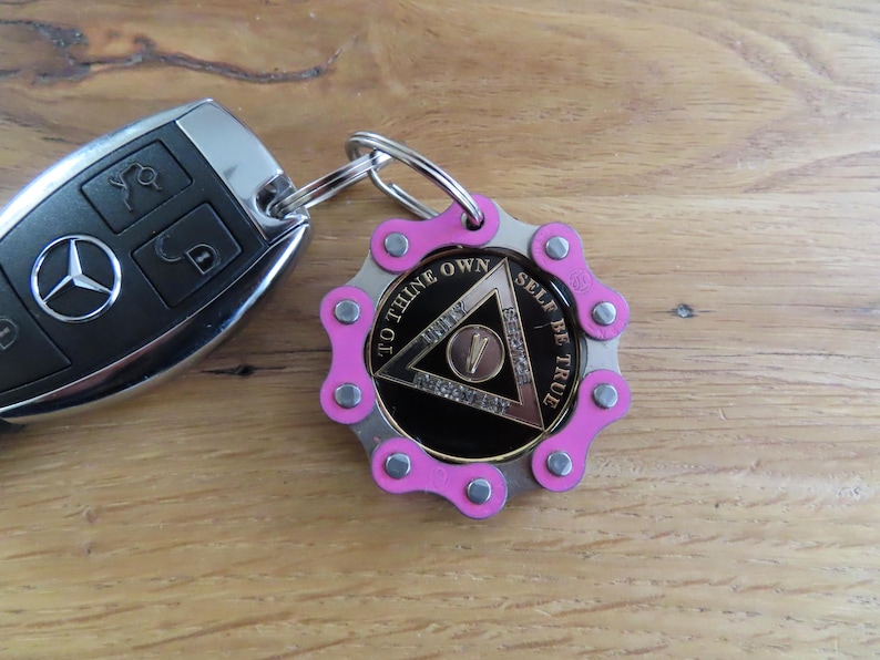 May include: A black Mercedes car key fob attached to a circular keychain. The keychain features a pink bicycle chain link border surrounding a black medallion with a silver triangle, the number five, and the text TO THINE OWN SELF BE TRUE.