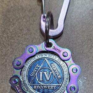 May include: A keychain featuring a light purple carabiner attached to a circular medallion. The medallion has an iridescent purple and blue bicycle chain border and a central blue disc with the Roman numeral IV, the words UNITY, SERVICE, RECOVERY, and TO THINE OWN SELF BE TRUE.