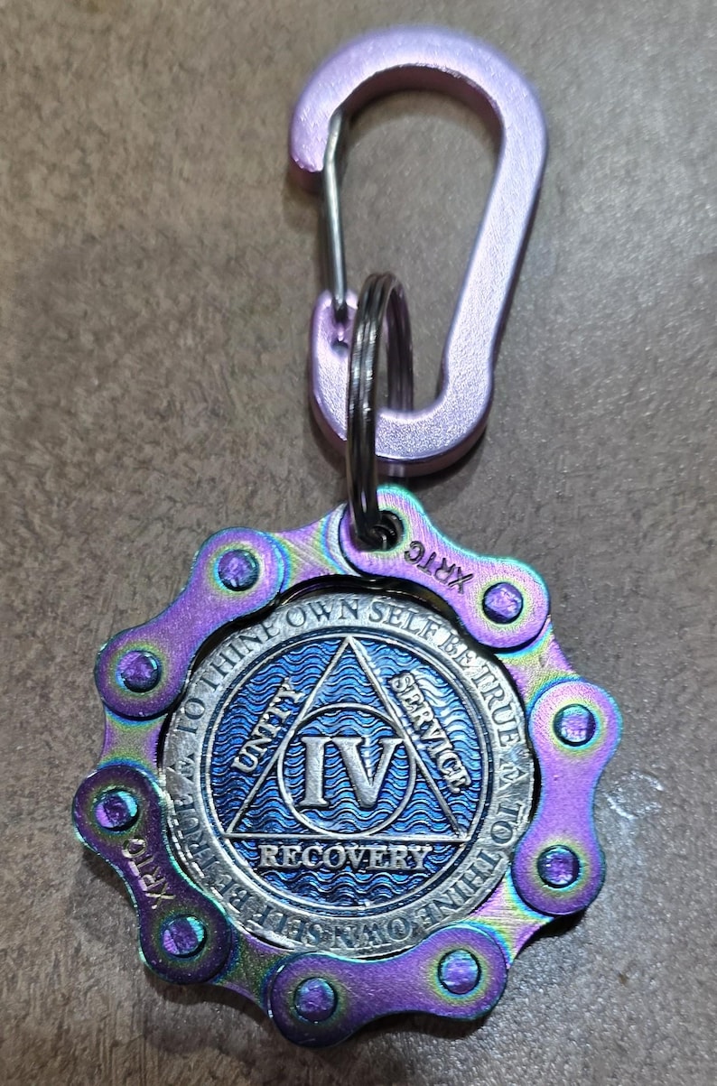 May include: A keychain featuring a light purple carabiner attached to a circular medallion. The medallion has an iridescent purple and blue bicycle chain border and a central blue disc with the Roman numeral IV, the words UNITY, SERVICE, RECOVERY, and TO THINE OWN SELF BE TRUE.
