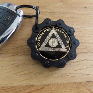 May include: A black metal keychain featuring a circular medallion with a gear-like border. The medallion displays a triangle with the text TO THINE OWN SELF BE TRUE, UNITY, SERVICE, RECOVERY, and a Roman numeral V in the center. Attached to a car key.