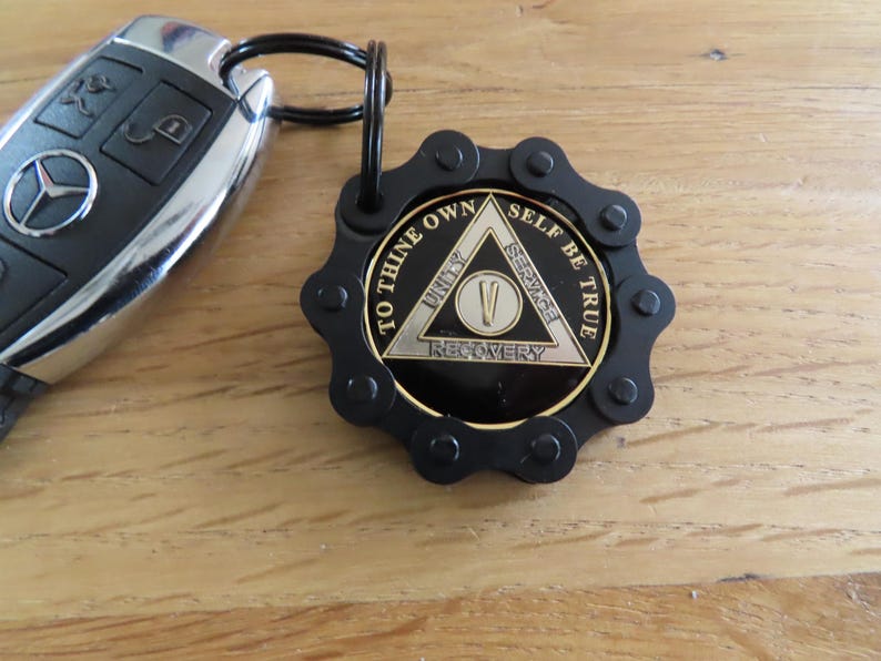May include: A black metal keychain featuring a circular medallion with a gear-like border. The medallion displays a triangle with the text TO THINE OWN SELF BE TRUE, UNITY, SERVICE, RECOVERY, and a Roman numeral V in the center. Attached to a car key.