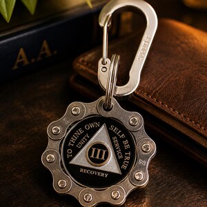 May include: A silver-toned metal keychain featuring a circular gear-like outer frame and a central black medallion. The medallion displays a silver triangle with the Roman numeral III, surrounded by the text TO THINE OWN SELF BE TRUE.