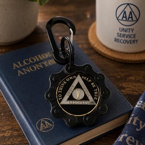 May include: A black metal keychain featuring a gear-shaped outer frame and a central triangular emblem. The emblem displays the Roman numeral V, the words Unity, Service, Recovery, and the phrase To Thine Own Self Be True in silver and black.