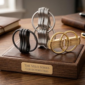 Può includere: Un espositore in legno presenta tre portachiavi in metallo: nero, argento e oro. Ogni portachiavi presenta un design unico con più segmenti ad incastro. L'espositore ha una placca dorata con la scritta "The Velo Series Collector's Trio."