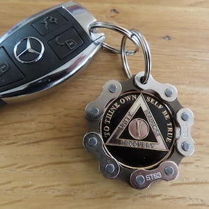 May include: A black Mercedes-Benz car key fob attached to a circular keychain. The keychain features a metallic bicycle chain link border surrounding a black medallion with a gold triangle, the Roman numeral V, and the text To Thine Own Self Be True.