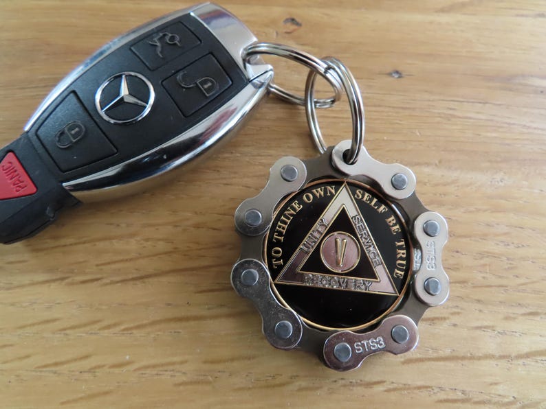 May include: A black Mercedes-Benz car key fob attached to a circular keychain. The keychain features a metallic bicycle chain link border surrounding a black medallion with a gold triangle, the Roman numeral V, and the text To Thine Own Self Be True.