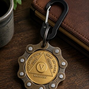 May include: A circular metal keychain featuring a brass-colored center medallion with a triangle, the Roman numeral V, and the text TO THINE OWN SELF BE TRUE, UNITY, SERVICE, RECOVERY, and XRTC, surrounded by a dark metallic bicycle chain link border.