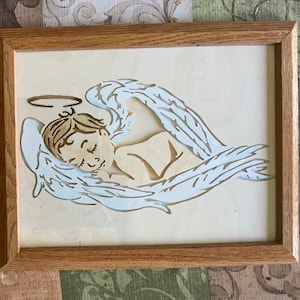 Puede incluir: Una obra de arte enmarcada en madera que representa un ángel durmiente con un halo, acunado en alas blancas. El pelo del ángel y el marco son marrones, sobre un fondo beige claro.