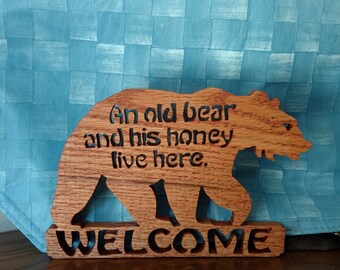 May include: Wooden bear-shaped welcome sign with the text "An old bear and his honey live here" and "WELCOME" cut out. The sign is made of brown wood and is placed on a dark wooden surface.