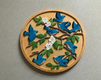May include: A round wooden wall art piece featuring bluebirds perched on branches with green leaves and white flowers. The birds are painted in shades of blue with orange accents, set against a natural wood background.