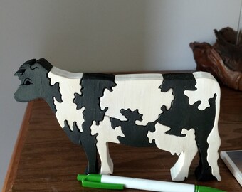 May include: A wooden cow figurine with a black and white pattern. The cow is standing on a wooden surface. A green and white pen is lying in front of the cow.