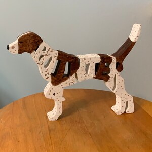 Può includere: Un puzzle di legno a forma di cane in posizione eretta. Il cane presenta sezioni marroni e bianche, con un motivo a pois sulle aree bianche. Il puzzle è assemblato e si trova su una superficie di legno.