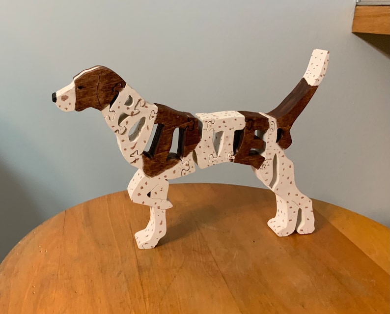 May include: A wooden dog puzzle, assembled and standing. The dog features brown and white sections with a spotted pattern on the white areas. It is positioned on a wooden surface. The puzzle is a decorative item.