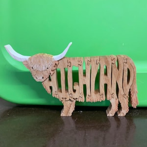 May include: Wooden Highland cow puzzle with the word "HIGHLAND" cut out of the body. The cow has white horns and a shaggy mane. The puzzle is made of light-colored wood and sits on a dark surface against a green background.