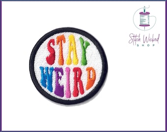 May include: A round, black-bordered patch with white fabric in the center. The words "STAY WEIRD" are embroidered in rainbow colors.