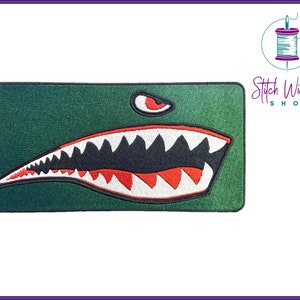 Puede incluir: Parche bordado verde con un diseño de dientes de tiburón blanco y rojo. El parche tiene un ojo negro con una pupila roja.
