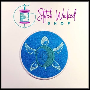 K&ouml;nnte beinhalten: Ein blauer gestickter Aufn&auml;her mit einem wei&szlig;en Umriss einer Meeresschildkr&ouml;te mit Fl&uuml;geln. Die Schildkr&ouml;te befindet sich auf einem blauen Hintergrund mit einem wei&szlig;en Kreis am Rand.