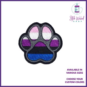 May include: A grey paw print embroidered with the colours of the genderqueer pride flag. The paw print is outlined in grey and has a pink, white, purple, black, and blue stripe pattern. The text "Stitch Wicked Shop" is in the top right corner of the image.