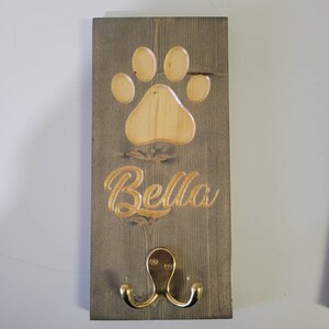 May include: A wooden dog leash hook with a paw print design and the name "Bella" engraved on it. The hook is painted gray and has a gold metal hook.
