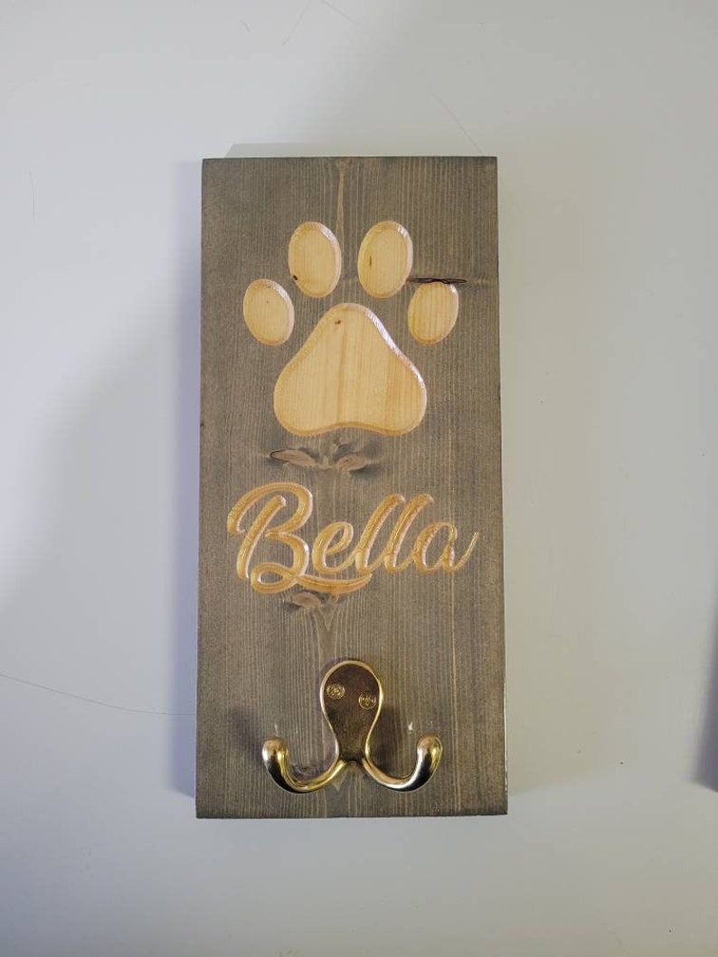 May include: A wooden dog leash hook with a paw print design and the name "Bella" engraved on it. The hook is painted gray and has a gold metal hook.