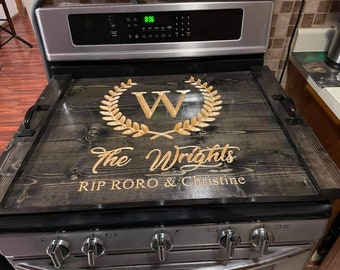 May include: A wooden stove cover with a dark gray finish. It features a carved gold-colored wreath with the letter "W" in the center, and the words "The Wrights" and "RIP RORO & Christine" are also carved into the wood. Black handles are attached.