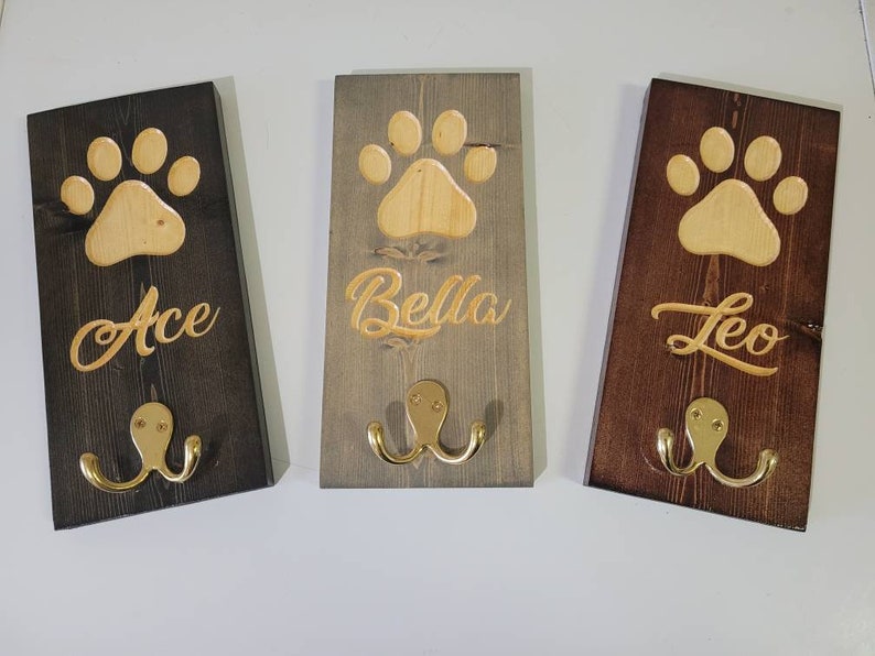 May include: Three wooden dog leash hooks with paw print designs and engraved names: Ace, Bella, and Leo. Each hook has two gold metal hooks.