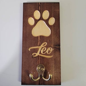 May include: A wooden dog leash hook with a paw print and the name "Leo" engraved on it. The hook is brown with a gold finish.