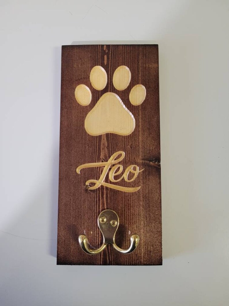 May include: A wooden dog leash hook with a paw print and the name "Leo" engraved on it. The hook is brown with a gold finish.