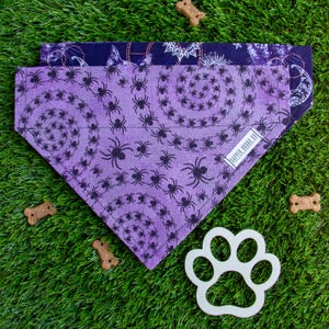May include: Two Halloween-themed dog bandanas. The top bandana is navy blue with a celestial pattern. The bottom bandana is purple with a spiral spider design. A white paw-shaped cutout and dog treats are also visible.