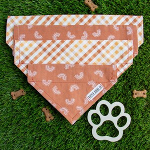 Op de afbeelding: Drie hondenbandana's in tinten oranje en wit, met regenboog- en ruitpatronen. De bandana's zijn gerangschikt op groen gras, met hondenkoekjes en een pootvormige uitsparing. De onderste bandana heeft de tekst "Rapper Doggo Co."