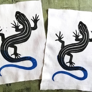 May include: Two white fabric patches each feature a black and blue lizard design. The lizard has a striped body and a curved blue tail. The patches are on a green surface.
