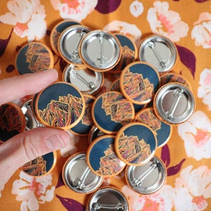 May include: A pile of round, orange and blue buttons with a mountain range design. The buttons have silver backs with safety pins.