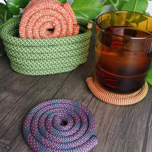 May include: A selection of handmade rope coasters and a basket. The coasters are in spiral patterns with red, orange, purple, and green colours. The basket is green and holds a red and green rope. A glass of amber liquid sits nearby.
