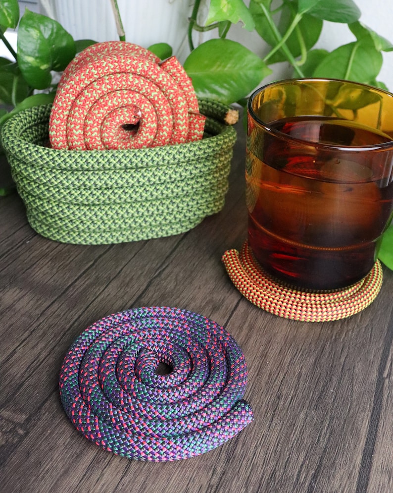May include: A selection of handmade rope coasters and a basket. The coasters are in spiral patterns with red, orange, purple, and green colours. The basket is green and holds a red and green rope. A glass of amber liquid sits nearby.