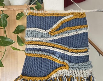 Pode incluir: Uma tapeçaria de parede tecida com um padrão geométrico azul, branco e amarelo. A tapeçaria tem uma borda de franjas e está montada em uma vareta de madeira.