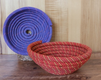 May include: A coiled purple rope and a red and yellow rope basket sit on a wooden surface. The coiled rope is circular, while the basket is oval-shaped. The basket is made of woven rope.