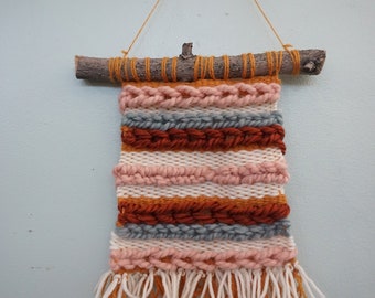May include: A woven wall hanging with a natural wood branch at the top. The hanging features a colourful striped pattern in shades of orange, pink, white, and grey. The bottom of the hanging has long, orange and white fringe.