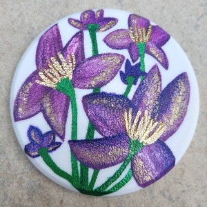 May include: A round, white ceramic coaster with a painted design of purple flowers with gold accents. The flowers have a textured look and are arranged in a cluster.